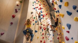 Four adults climbing a colorful indoor wall in a modern recreation center, showcasing teamwork and athletic challenge under bright lighting.