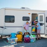 A couple loads colorful beach gear into storage bins inside an RV parked on a sunny, gravel lot with coastal grasses and sand dunes in the background.