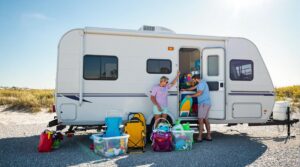 A couple loads colorful beach gear into storage bins inside an RV parked on a sunny, gravel lot with coastal grasses and sand dunes in the background.